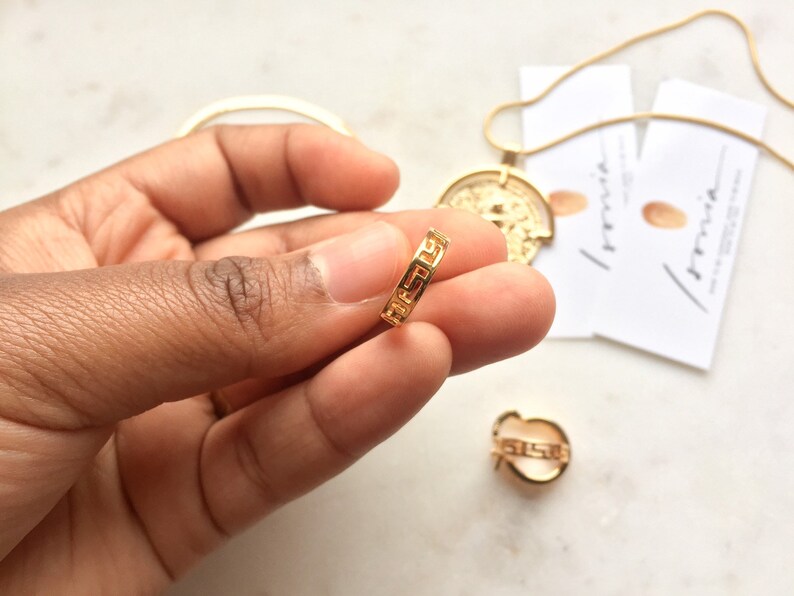 May include: Gold ring with a Greek key design. The ring is being held by a hand.