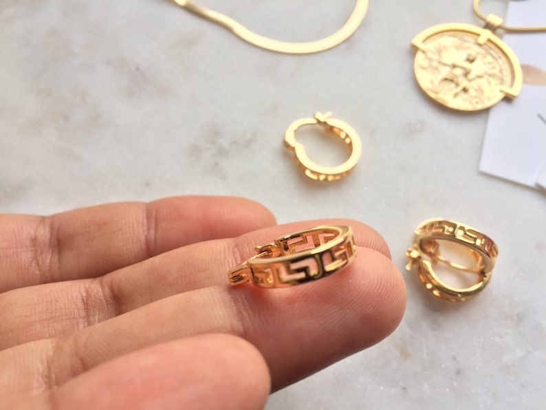 May include: Gold hoop earrings with a Greek key design. The earrings are shown on a white surface.