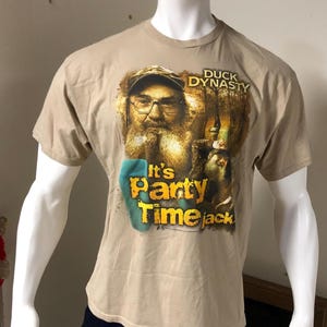 Könnte beinhalten: Beiges T-Shirt mit einem Aufdruck eines Mannes mit langem Bart und Brille, mit dem Text "Duck Dynasty" und "It's Party Time Jack". Das T-Shirt wird auf einer Schaufensterpuppe präsentiert.