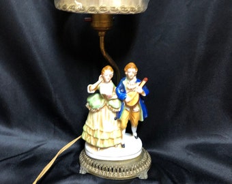 Può includere: Una lampada da tavolo in porcellana con una base dorata e un paralume color crema. La base presenta una statuetta di una coppia, una donna con un vestito verde e giallo e un uomo con una giacca blu che suona uno strumento a corda.