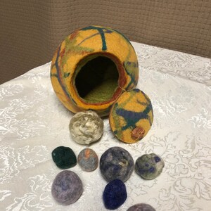 May include: A yellow, green, and blue felted wool ball with a lid, and a collection of smaller felted balls in various colors, including green, purple, blue, and gray.
