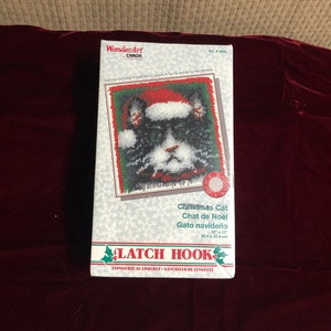 Peut inclure: Un kit de crochet à boucle pour réaliser un chat noir et blanc portant un bonnet de Père Noël rouge. Le kit est intitulé "Chat de Noël" et comprend des instructions pour créer un projet de crochet à boucle de 12 x 12 pouces (30,4 x 30,4 cm).