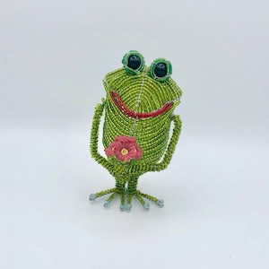 以下が含まれることがあります： ピンクの花を持った緑色のビーズでできたカエルの置物。カエルは黒い目と赤い笑顔をしています。