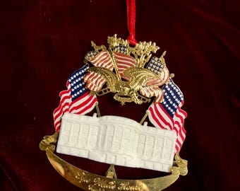 Puede incluir: Adorno navideño dorado con la Casa Blanca, banderas americanas y un águila. El adorno tiene una cinta roja para colgar. El texto en el adorno dice "The White House, Washington 1988."