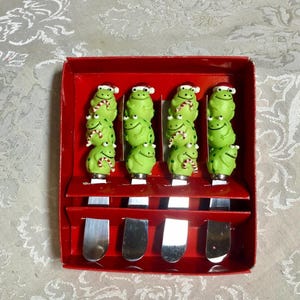 May include: A set of four cheese spreaders in a red box. Each spreader has a silver blade and a green frog handle wearing a Santa hat and a candy cane scarf. The frogs are stacked on top of each other.