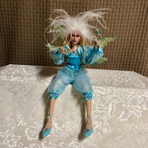 May include: A handcrafted fairy doll with a blue and turquoise color scheme. The doll has a feathered headdress, blonde hair, and delicate wings. She wears a blue bodice, puffy pants, and matching shoes. The doll is seated on a patterned surface.