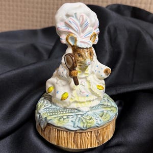 Puede incluir: Figura de cerámica de un ratón con gorro y vestido, sosteniendo un pequeño espejo. El ratón está sobre una base redonda con efecto de madera y diseño floral. La figura está pintada en colores pastel.