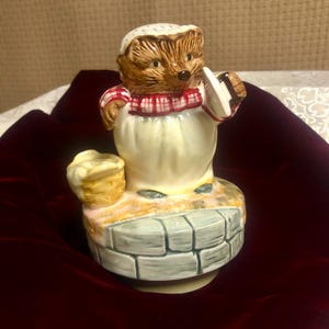 Peut inclure: Figurine en céramique représentant un hérisson portant un bonnet blanc et une écharpe à carreaux rouges et blancs. Le hérisson tient une assiette et se tient sur un puits. Un panier de pain est posé à côté du puits.