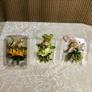 May include: Three miniature fairy figurines. One is encased in a clear plastic box and dressed in a yellow flower costume. The other two are free-standing, one in green and white, and the other in pink and green.