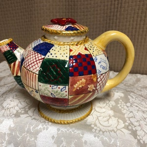 May include: A ceramic teapot with a patchwork design in shades of red, blue, green, and yellow. The teapot has a gold trim and a red flower on the lid.
