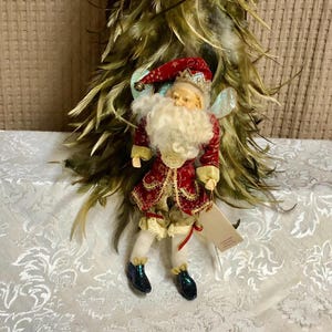 May include: A decorative figurine of a fairy dressed in a red and gold coat with a white beard and a red hat. The fairy has blue wings and black shoes. A feather decoration is behind the fairy.