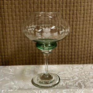 May include: A clear glass candle holder with a green base and stem. The bowl-shaped top has an etched design of a heart, crown, and hands. The candle holder sits on a round base and is displayed on a white patterned surface.