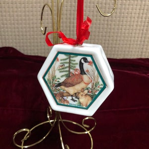 Könnte beinhalten: Weißer, sechseckiger Weihnachtsbaumschmuck aus Porzellan mit rotem Band. Der Anhänger zeigt ein gemaltes Bild einer Gans, eines Kaninchens und eines kleinen Vogels in einer Winterwaldlandschaft. Der Text "The Power of Giving" ist auf der Unterseite des Anhängers aufgedruckt.