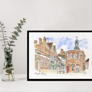 May include: Watercolor painting of a town square with brick buildings, a clock tower, and a blue sky. The artwork is framed in a black frame and includes the text "Leigh Surrey". A vase with green leaves is in the foreground.