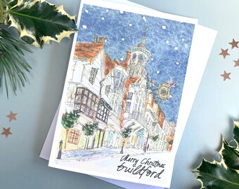 以下が含まれることがあります： イギリスのギルフォードの街並みを水彩画で描いたクリスマスカード。雪が降る夜に、時計塔とクリスマスのイルミネーションで飾られた建物が描かれている。カードの下部には「メリークリスマス ギルフォード」という文字が筆記体で書かれている。