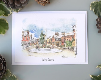 以下が含まれることがあります： 雪の降る街の広場とクリスマスツリーを描いた水彩画のクリスマスカード。「Merry Christmas」と地名「Haslemere」の文字が入っています。松ぼっくりと緑に囲まれたカードです。