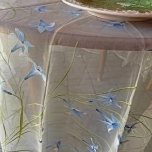 May include: A sheer, light-colored tablecloth with a delicate floral pattern. The design features blue bell-shaped flowers and green stems, creating an elegant and airy aesthetic. The tablecloth is draped over a wooden table.