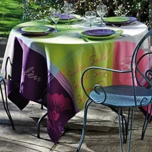 May include: A round, white tablecloth with a colourful design featuring pink, purple, green, and yellow. The tablecloth has the words "Soleil" and "Soleil Solaire" printed on it. The tablecloth is set on a table with four chairs, two of which are visible in the image. The chairs are made of black metal and have a blue finish.
