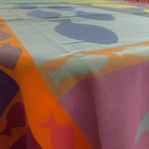Peut inclure: Une nappe rectangulaire présentant un motif de poissons répété et disposé en blocs de couleurs distinctes. Le design comprend des nuances d'orange, de violet, de bleu clair et de jaune, avec des silhouettes de poissons imprimées sur le tissu.
