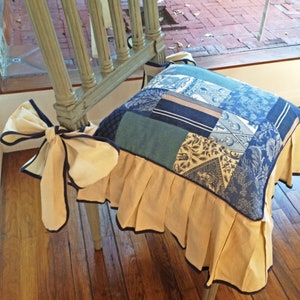 May include: A chair cushion with a patchwork design in shades of blue and beige. The cushion has a ruffled edge and a blue ribbon tied around the back.
