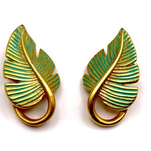 May include: A pair of gold-toned leaf-shaped earrings with teal accents. The earrings have a textured, ribbed design and a curved, looped base. The earrings are set against a white background.