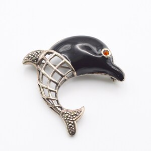 Peut inclure: Une broche en argent en forme de dauphin avec un émail noir et un œil en strass rouge. La queue du dauphin est en marcassite.