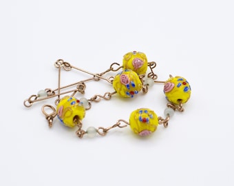 May include: A yellow glass beaded necklace with floral designs in pink, blue and red. The beads are connected by a thin gold chain.