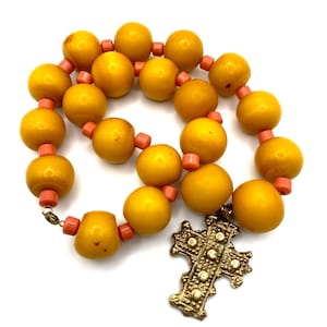 Puede incluir: Un collar de cuentas de ámbar amarillo con un colgante de cruz dorada. Las cuentas están separadas por pequeñas cuentas de color coral.