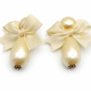 Puede incluir: Dos adornos perlados en forma de ángel con lazos color crema. Los adornos tienen forma de lágrima con un pequeño elemento floral decorativo en la base. El fondo es blanco.