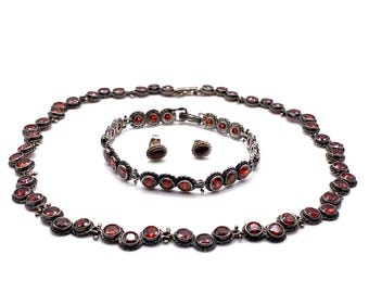 Puede incluir: Un conjunto de joyas con collar, pulsera y anillo. El conjunto tiene metal plateado con piedras preciosas rojas y redondas. El collar y la pulsera están unidos. El anillo tiene una sola piedra preciosa.