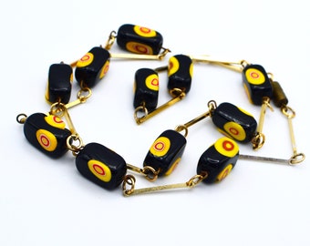 May include: A gold chain necklace with black and yellow rectangular beads. Each bead has a red and yellow circle design.
