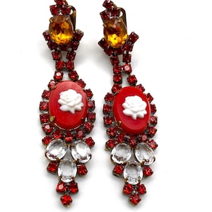 Peut inclure: Une paire de boucles d'oreilles vintage à clip. Chaque boucle d'oreille présente un ovale rouge avec un camée de rose blanche, entouré de strass rouges. Des cristaux clairs en forme de goutte et des pierres ambrées complètent le design.