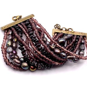 May include: A multi-strand bracelet featuring a mix of purple beads and iridescent pearls. The bracelet has a gold-tone clasp and a decorative gold-tone bar. The bracelet is a fashion accessory.