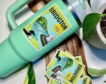 Könnte beinhalten: Eine grüne Reise-Thermoskanne mit Henkel und Strohhalm. Die Kanne hat einen Aufkleber mit der Aufschrift "Bridgton Maine" und zeigt einen Seetaucher, einen See und eine Straße. Auf einer Holzoberfläche befinden sich drei weitere Aufkleber.