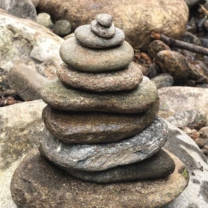 May include: A stack of gray and brown stones balanced on top of each other in a pyramid shape. The stones are arranged in a natural setting with other rocks and a stream in the background.