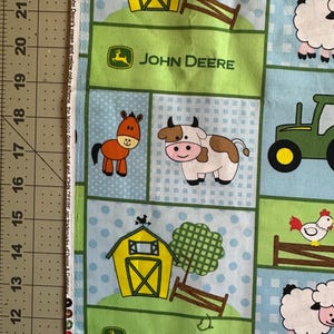 May include: Fabric with a repeating pattern featuring farm animals, barns, and tractors. The design includes a horse, cow, sheep, and chicken, with the John Deere logo and text. The background is a mix of green and light blue squares.