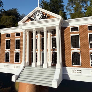 Pode incluir: Um modelo detalhado de um edifício de tijolo com colunas brancas e uma torre de relógio. O edifício tem uma fachada branca com uma escada cinza que leva à entrada. O mostrador do relógio é preto com ponteiros brancos e algarismos romanos.