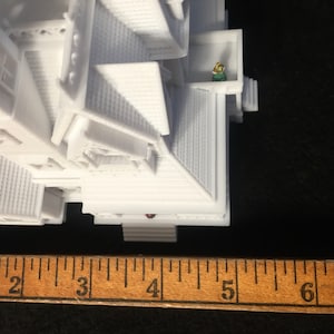 May include: White 3D printed model of a house with a small figure standing on a balcony. The house is approximately 4 inches tall.