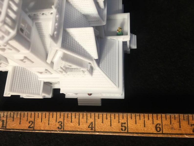 May include: White 3D printed model of a house with a small figure standing on a balcony. The house is approximately 4 inches tall.