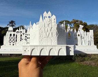 Könnte beinhalten: Ein weißes 3D-gedrucktes Modell des ikonischen Disney World Schlosses mit filigranen Details und einem verspielten Design. Das Schloss ist von kleineren Gebäuden umgeben, die eine Miniatur-Stadtlandschaft bilden.