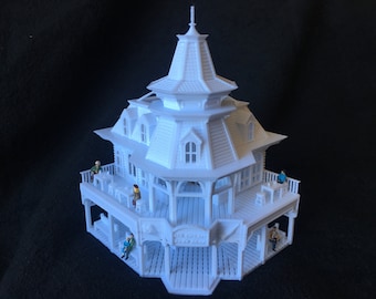 May include: White 3D printed model of a Victorian-style house with a wraparound porch and multiple levels. The house has a steep roof with a pointed spire and decorative details. There are small figures of people sitting on the porch.