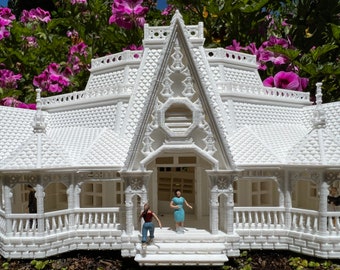May include: A detailed white miniature house with intricate architectural details, including a porch, balcony, and decorative trim. Small figurines are positioned on the steps and porch. The house is set against a backdrop of vibrant purple flowers.
