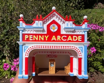 May include: A miniature model of a red, white, and blue Penny Arcade building with a sign that reads "PENNY ARCADE". The building has a white awning with red polka dots and a small window with a sign that reads "CANDY".