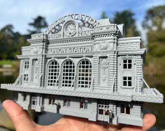 May include: A gray 3D printed model of a train station with the words "Union Station" and "Travel" on the front. The model is detailed with windows, doors, and a clock tower.