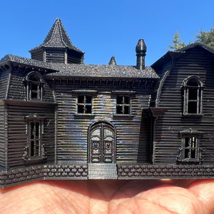 May include: A black 3D printed model of a haunted house with a pointed roof, a chimney, and multiple windows. The house is detailed with brickwork and siding.