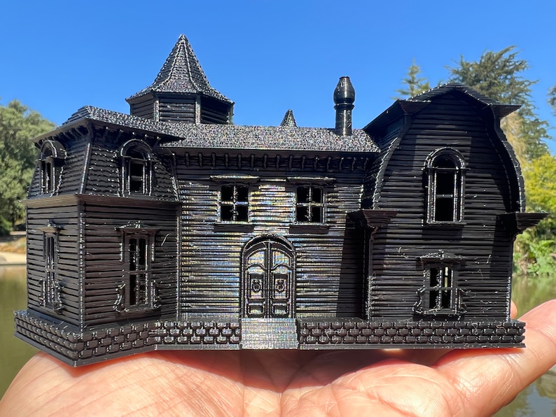 May include: A black 3D printed model of a haunted house with a pointed roof, a chimney, and multiple windows. The house is detailed with brickwork and siding.
