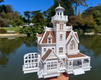 Op de afbeelding: Een gedetailleerd model van een Victoriaans huis in wit en bruin met een toren, een veranda en een kas. Het model is geplaatst tegen een wazige achtergrond van een vijver en bomen.
