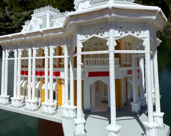 May include: A detailed, white miniature gazebo with intricate architectural details, including columns, arches, and decorative trim. The model features red and yellow accents and is held against a backdrop of water and trees.