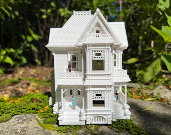 Puede incluir: Un modelo de casa victoriana impreso en 3D en blanco con una fachada detallada. La casa tiene un porche con escalones y un balcón en el segundo piso. Hay figuras en miniatura de pie en el porche y el balcón.