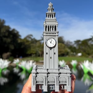 May include: A gray miniature replica of a clock tower with a clock face showing the time. The tower has architectural details and is held in front of a blurred background of a pond and trees.